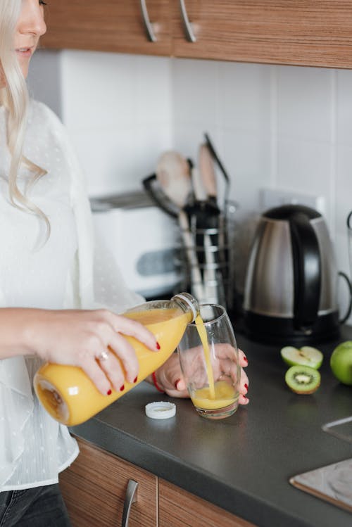 Free Crop unrecognizable lady in casual outfit pouring fresh orange juice in glass while standing in light kitchen near counter with kiwi and apple near kitchenware Stock Photo