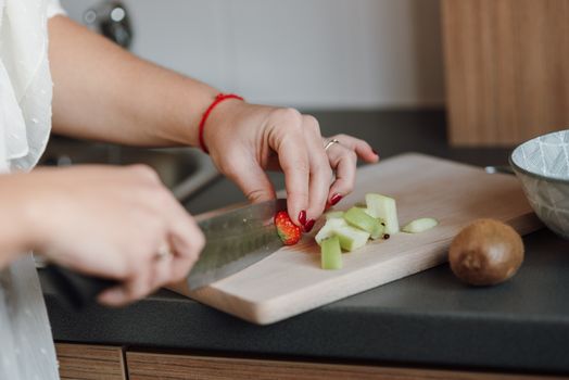 Crop anonymous woman cutting fresh strawberry on wooden cutting board near sliced apple while cooking healthy fruit salad in kitchen