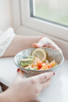 From above of crop anonymous woman in white clothes putting fresh fruit salad with kiwi and lemon on windowsill at home in daytime