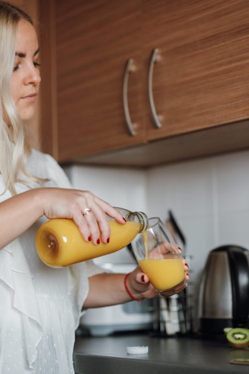 Free Crop young concentrated female pouring fresh orange juice from bottle into glass while standing in kitchen Stock Photo