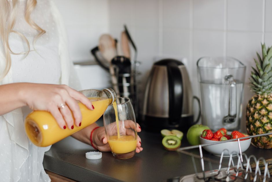 Crop faceless female standing near table with ripe fresh fruits in kitchen while pouring refreshing orange juice from bottle into glass