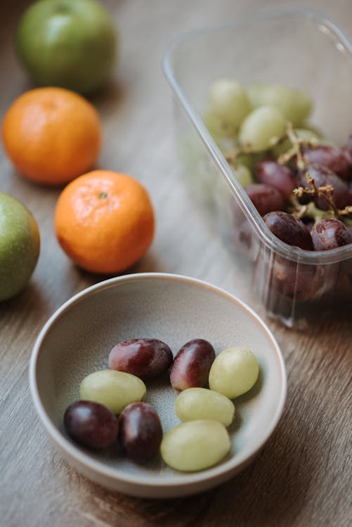 From above of green and red grapes in white bowl mandarins and green apples on wooden table