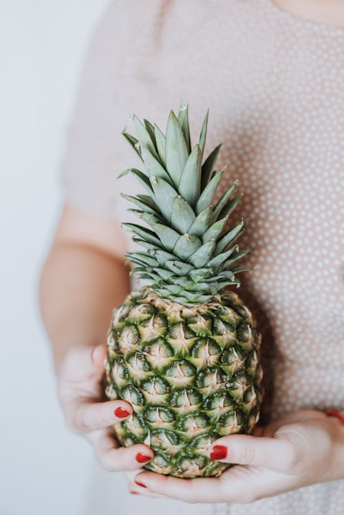 Unrecognizable woman in pink dress holding big ripe pineapple on grey background