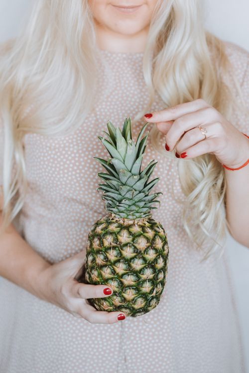 Free Unrecognizable long haired blond woman in pink dress holding ripe pineapple and touching spiky leaves Stock Photo