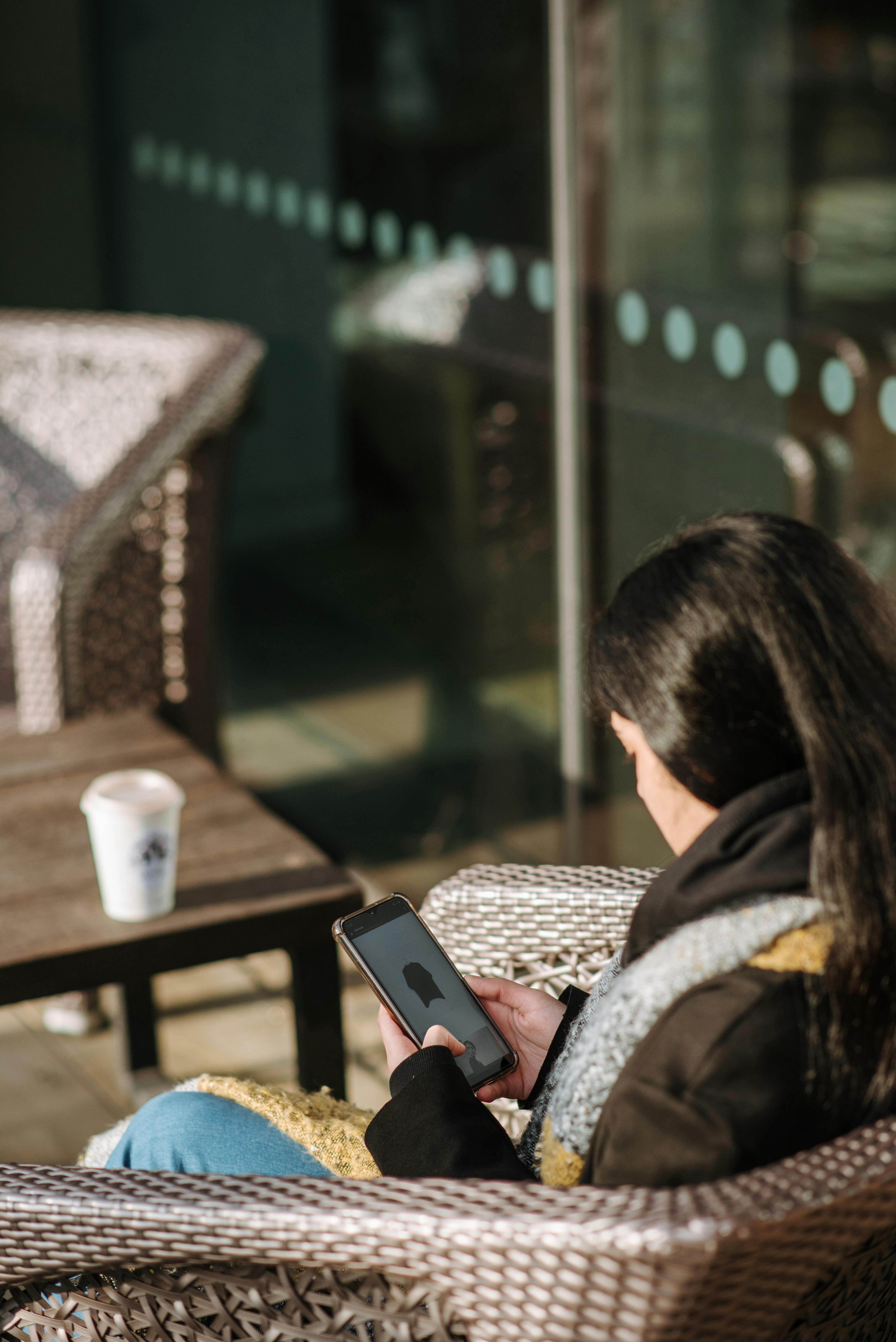 woman using smartphone while drinking hot beverage on cafe terrace