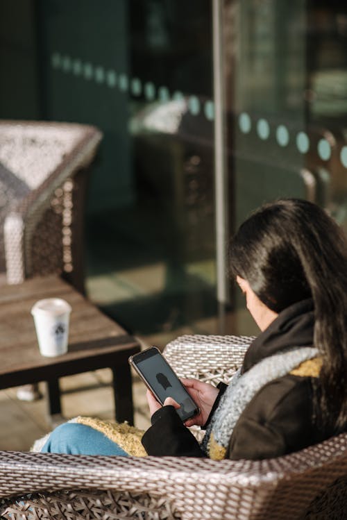 Free Side view of young female having rest in armchair and browsing mobile phone sitting in outdoor cafe Stock Photo
