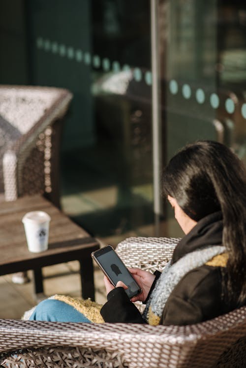 Side view of young female having rest in armchair and browsing mobile phone sitting in outdoor cafe