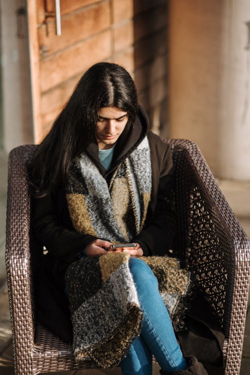 High angle of young ethnic female in warm clothes sitting in armchair on terrace and surfing internet on mobile phone