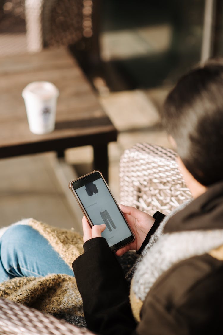 Young Woman Choosing Clothes In Internet Shop Sitting In Armchair In Cafeteria