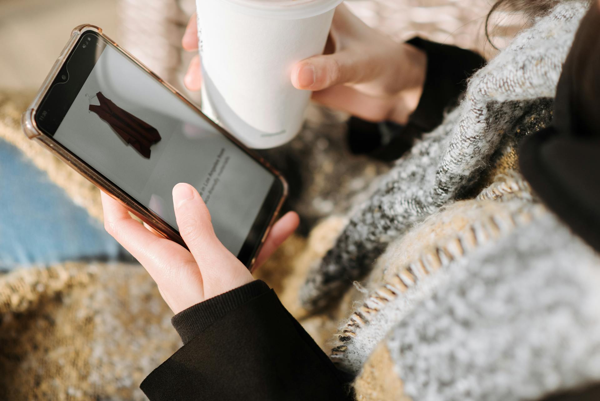 From above of crop anonymous female purchaser with takeaway hot drink choosing dress on cellphone screen during online shopping