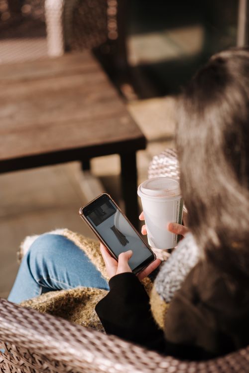 Free Back view of crop anonymous female purchaser choosing dress on mobile phone screen while sitting in armchair with beverage to go Stock Photo