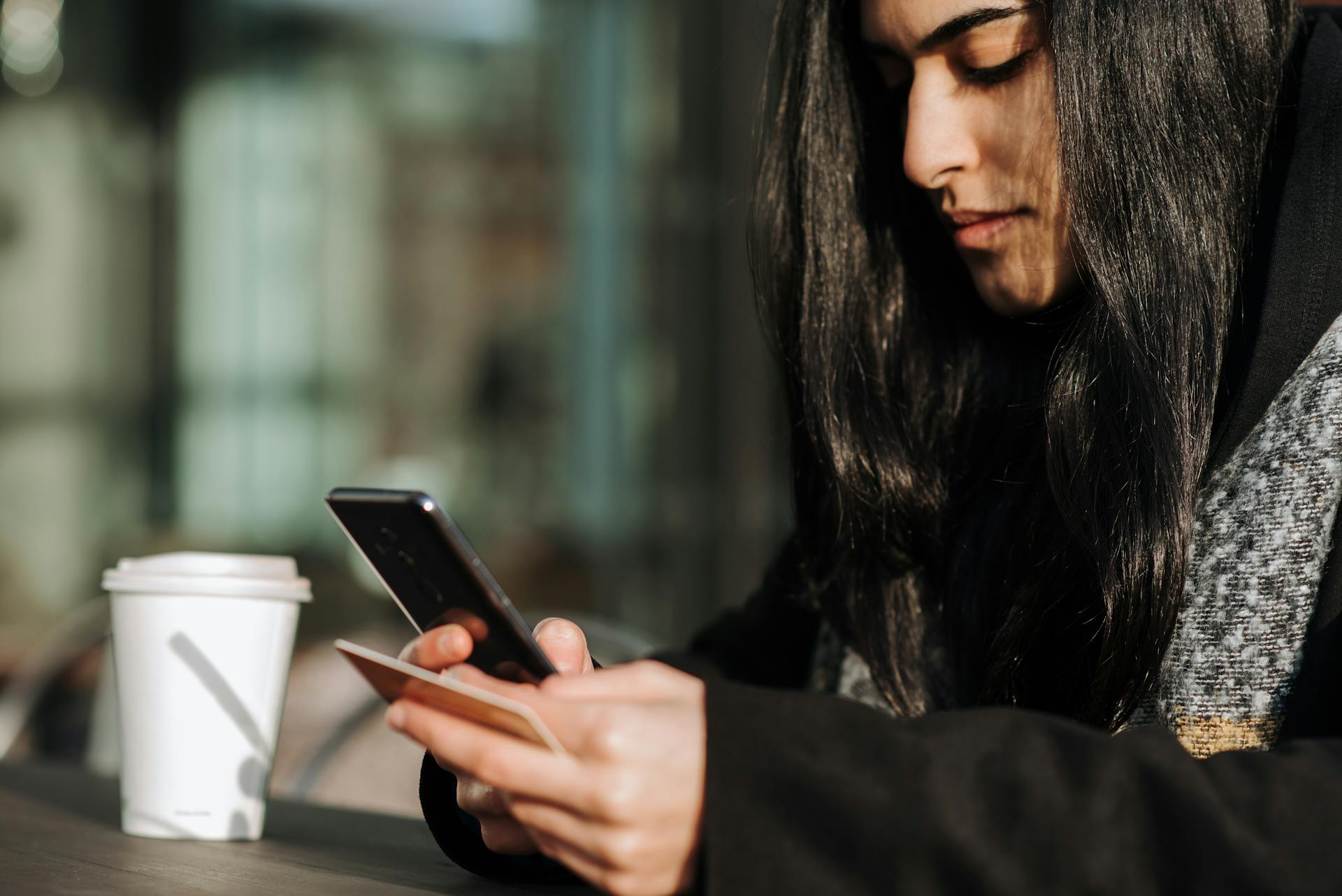 A young woman enjoys her coffee while making an online purchase using her smartphone and credit card.