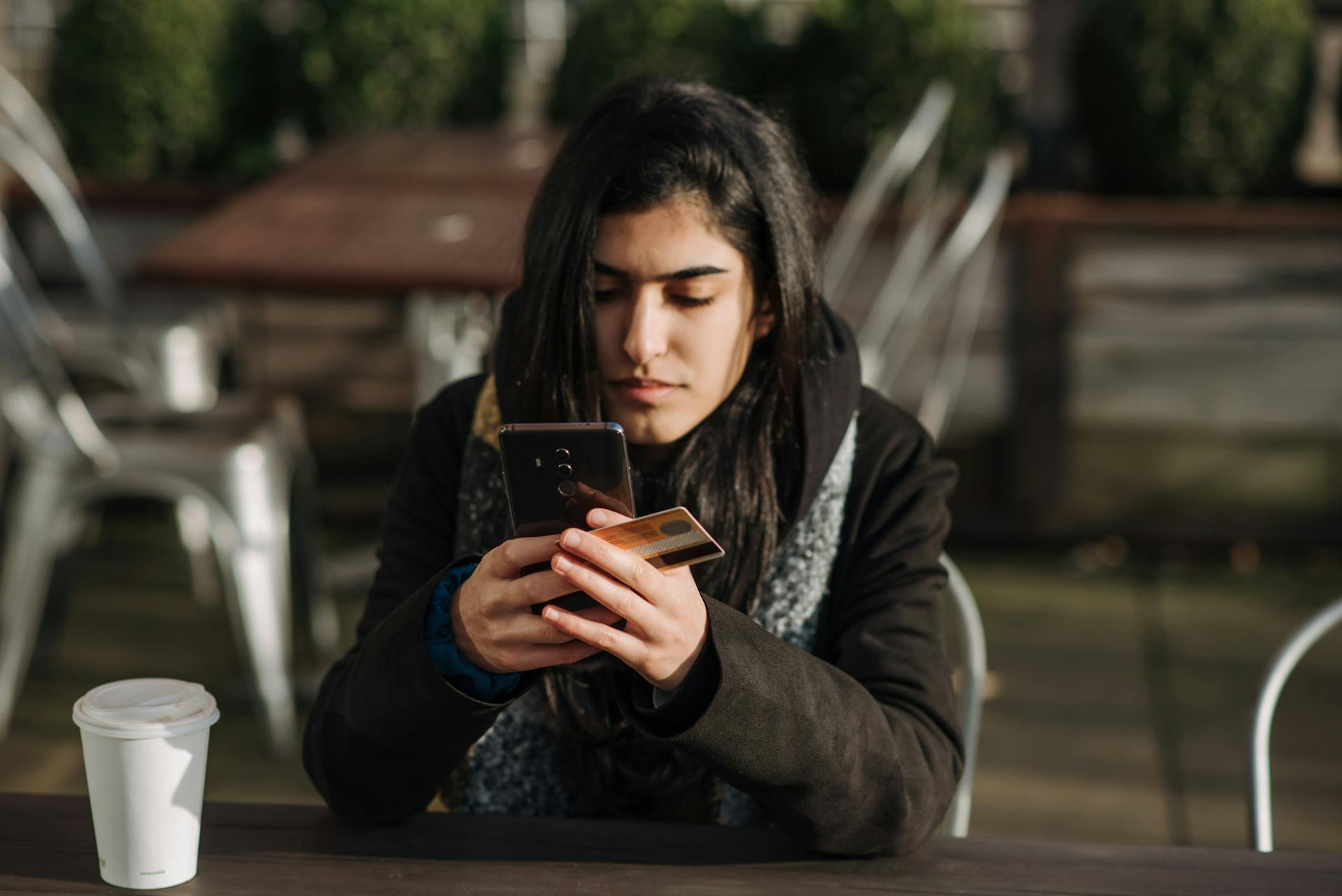 Young ethnic female purchaser with credit card shopping online on mobile phone at table with takeaway hot drink in cafeteria