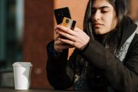 Crop young Hispanic female shopper with debit card making purchase on cellphone near takeaway beverage in cafeteria
