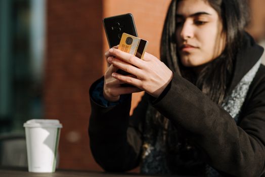 Crop young Hispanic female shopper with debit card making purchase on cellphone near takeaway beverage in cafeteria