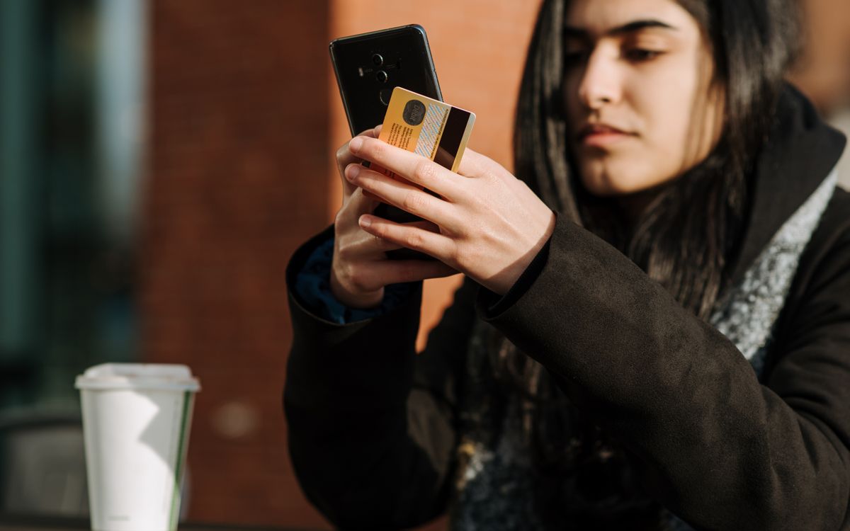 Crop young Hispanic female shopper with debit card making purchase on cellphone near takeaway beverage in cafeteria