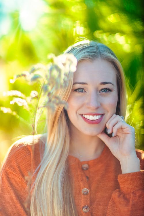 Cheerful pretty blonde wearing cozy red sweater touching face and looking at camera with happy smile while spending free time in sunny verdant nature