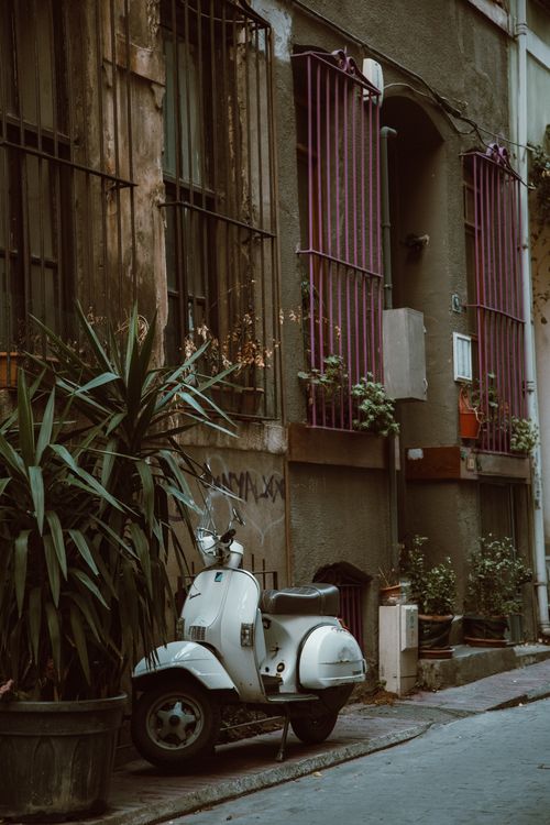 Free A vintage white scooter parked by a rustic urban street with potted plants and colorful window bars. Stock Photo