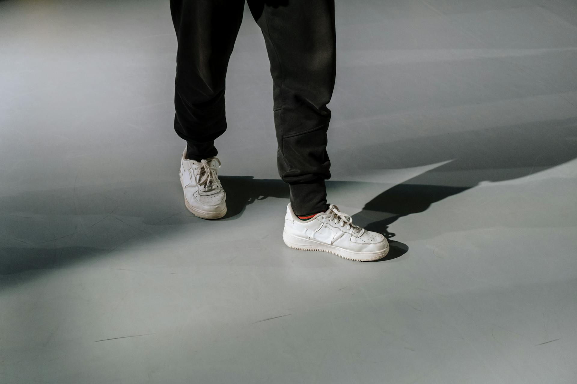 Person wearing white sneakers and black pants walking on a grey floor with shadow contrast.