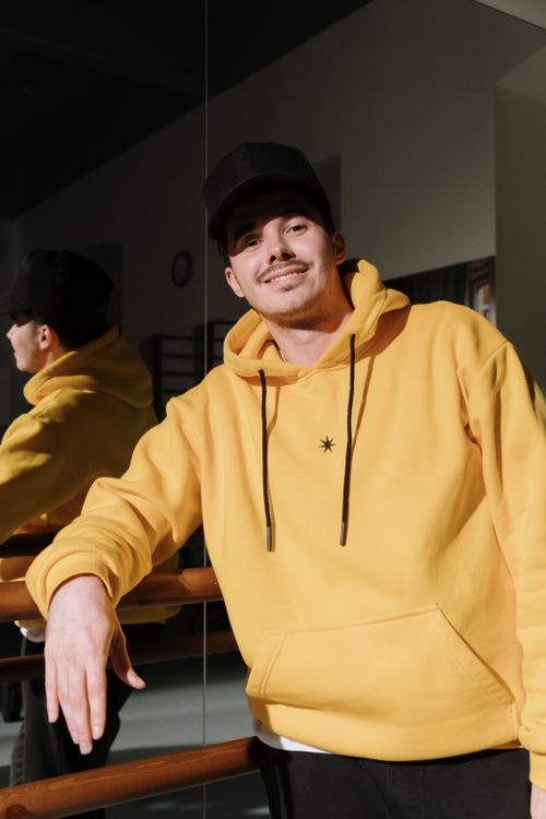 Free A man in a yellow hoodie smiles near a mirror indoors, adding warmth and style. Stock Photo
