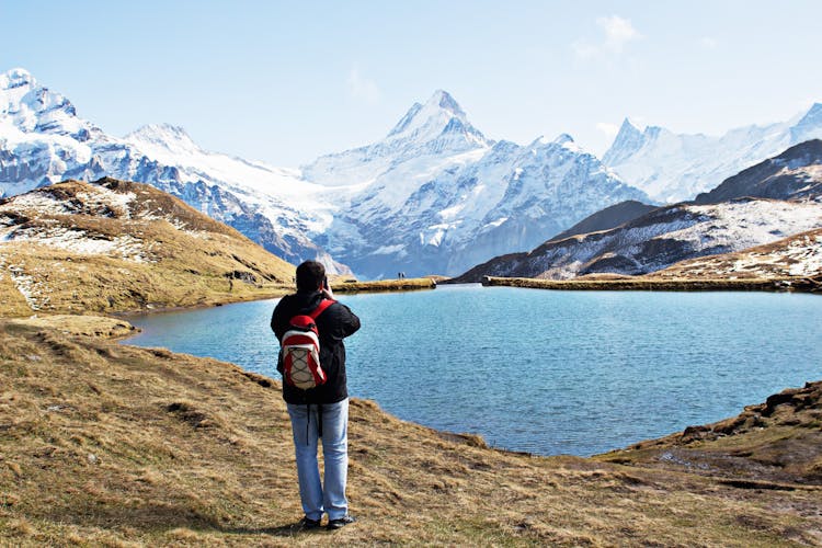 Standing Man Wearing White And Red Backpack In Front Of Calm Body Of Water