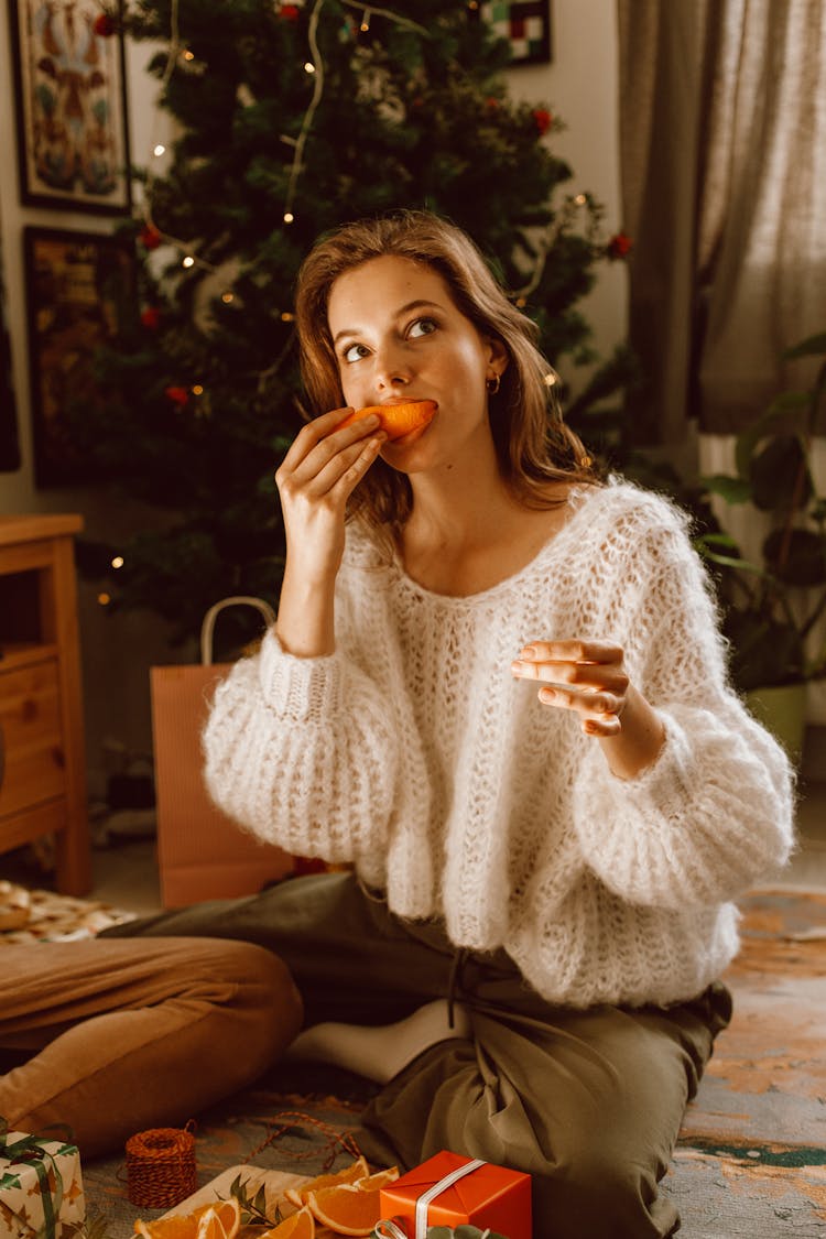 A Woman In White Sweater Sitting On The Floor While Eating Orange