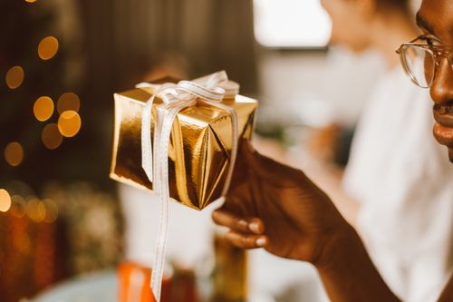 Free Close-up of a gold gift box with white ribbon held by a person, surrounded by warm bokeh lights. Stock Photo