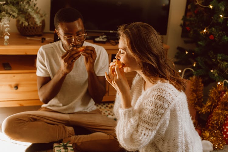 A Man And Woman Sitting On The Floor While Eating Oranges