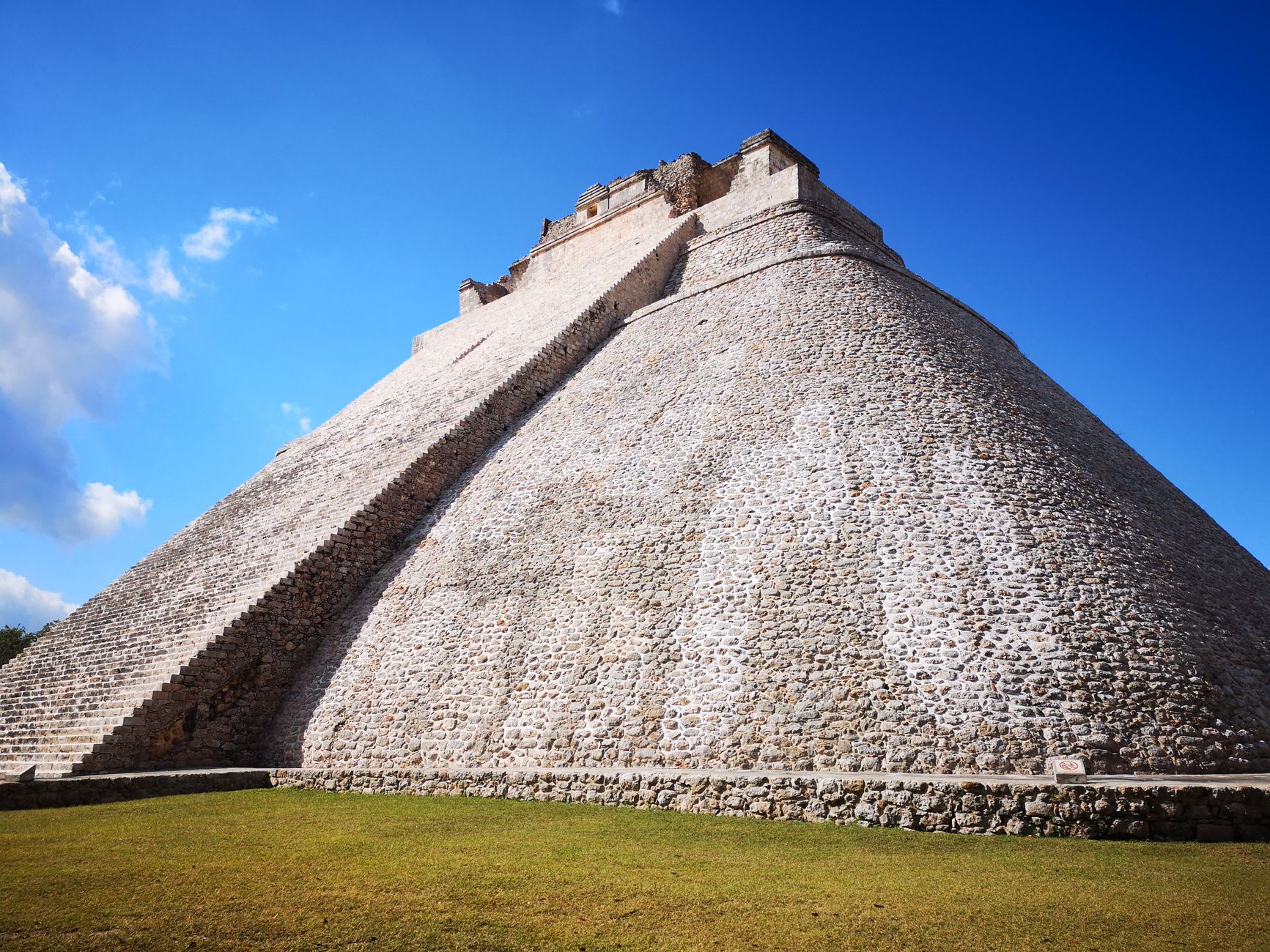 The tall, rounded-corner Pyramid of the Magician at the Maya ruins of Uxmal, its steep grey-stone flank sweeping up to the temple against a vivid blue sky