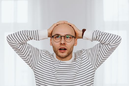 Bald man in striped shirt holding head in surprise indoors.