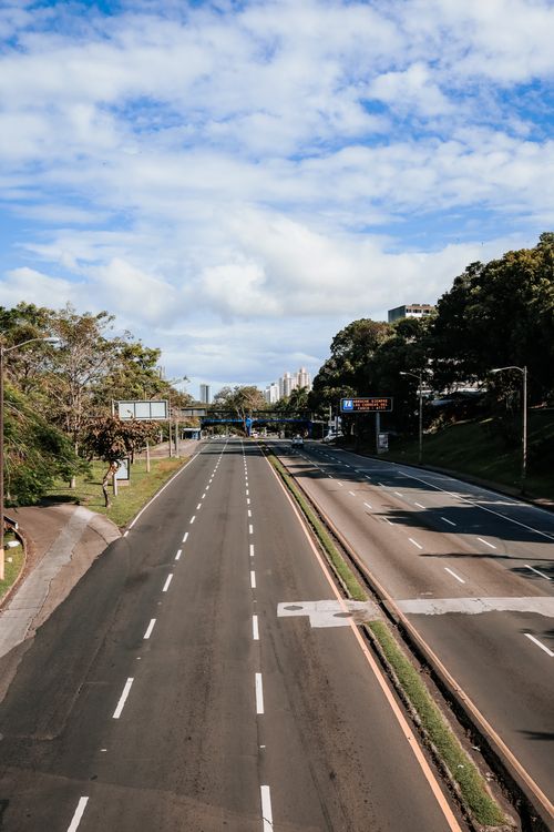 Free A wide, empty urban highway photographed on a clear, bright day, with lush greenery along the roadside and blue skies overhead. Stock Photo