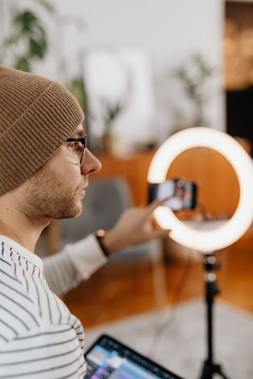 Free A man wearing glasses and a knit hat uses a smartphone with a ring light indoors. Stock Photo