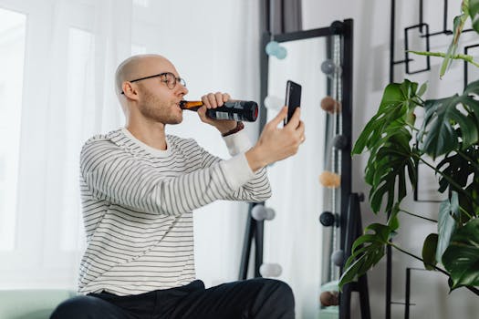 Bald man holding smartphone, drinking beer, enjoying a video call indoors.