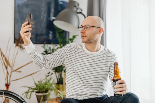 A Man Wearing Eyeglasses while Holding a Beer Bottle and His Phone