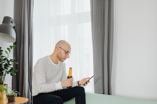 Free A Bald Man Drinking Beer While Holding a Smartphone Stock Photo