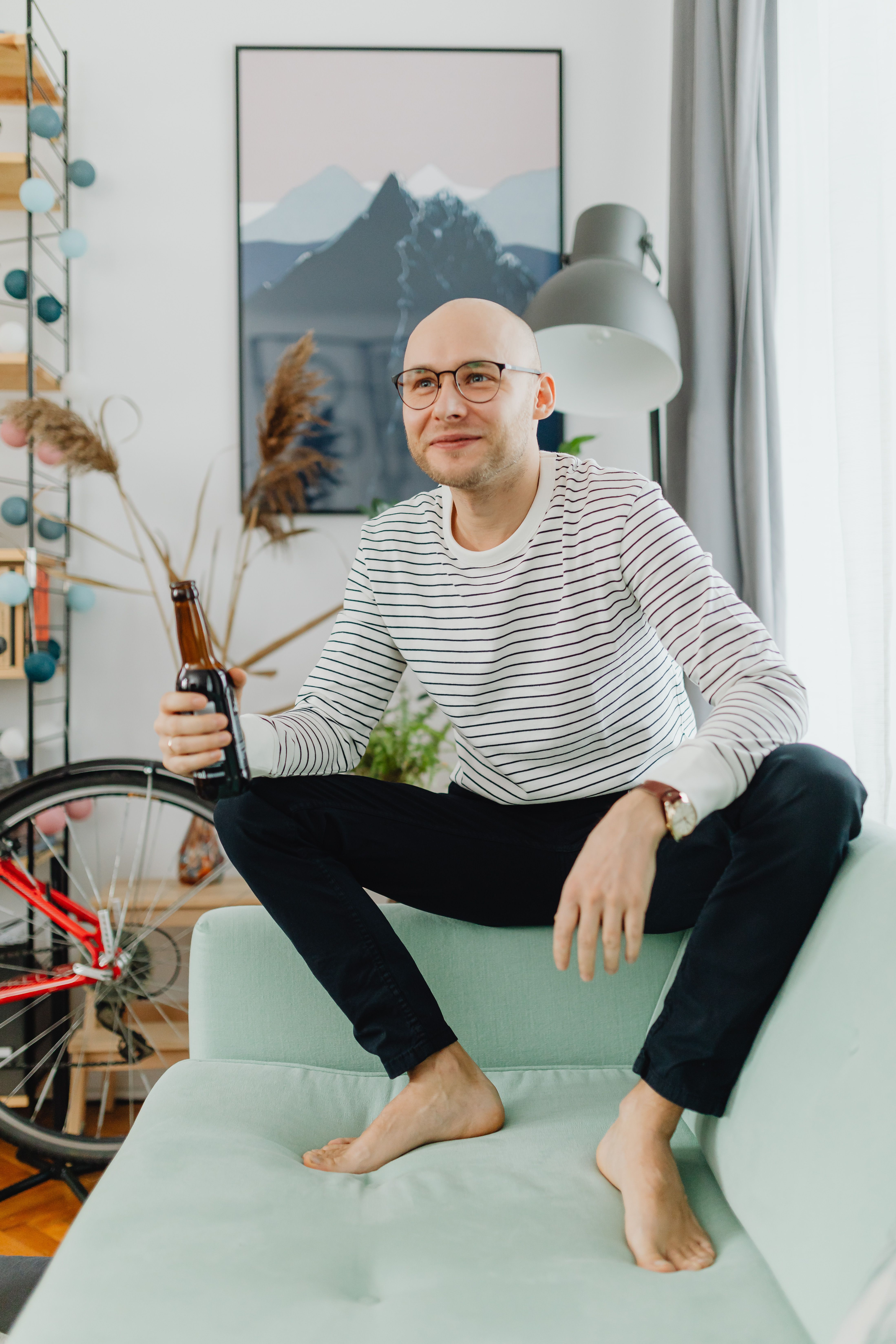 Bald man sitting on a couch holding a beer bottle in a cozy living room setting.