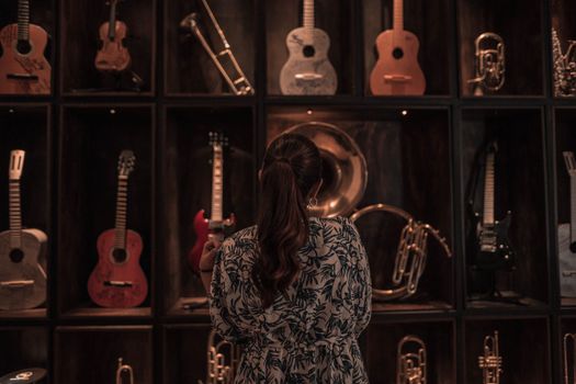 A woman observes a diverse collection of musical instruments on display, including guitars and trumpets, in a dimly lit room.