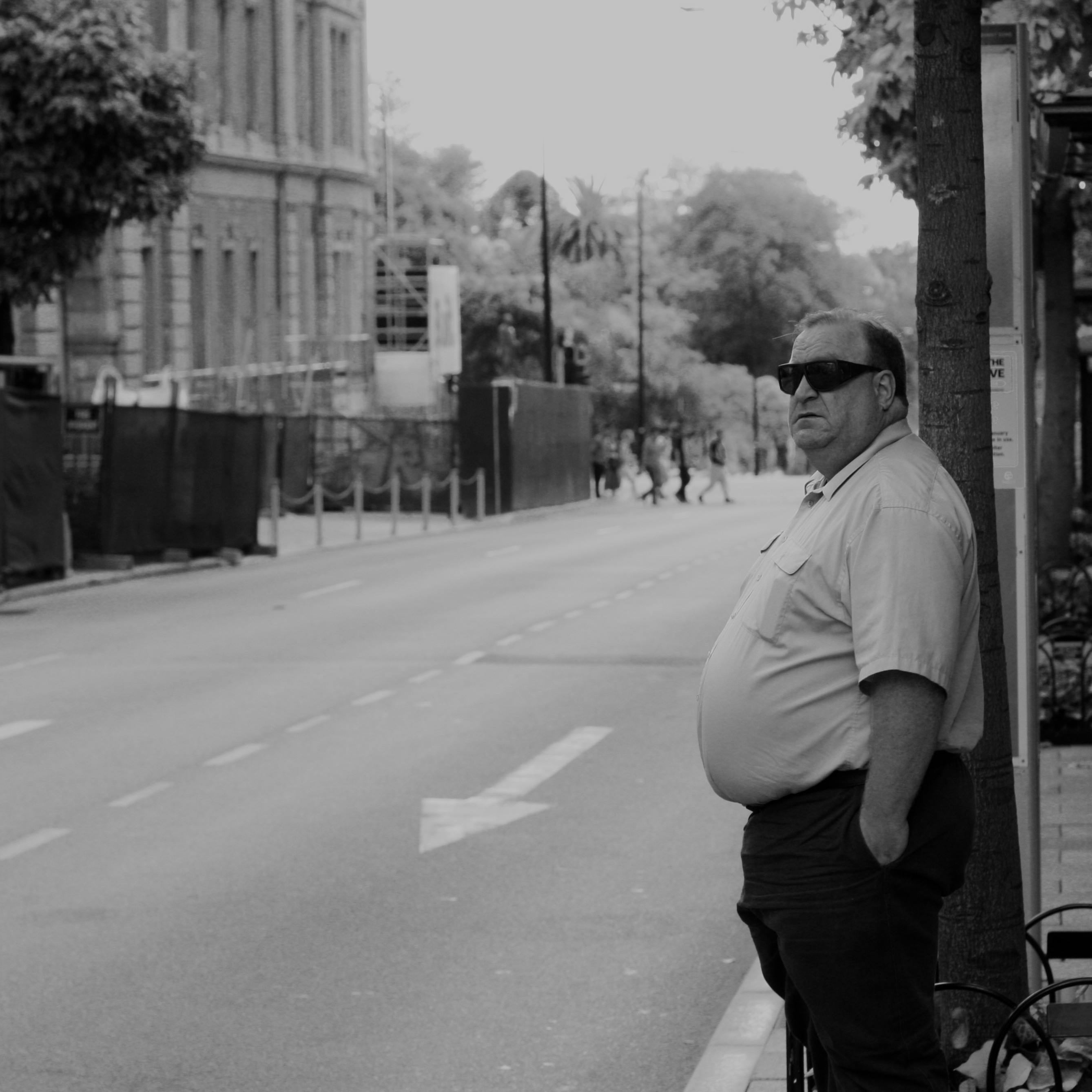 A man in sunglasses stands by an empty street in Perth, captured in black and white, showcasing urban life.