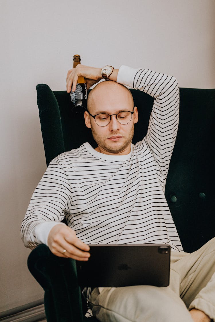 A Man Watching On His Ipad While His Hand On His Head With A Glass Bottle