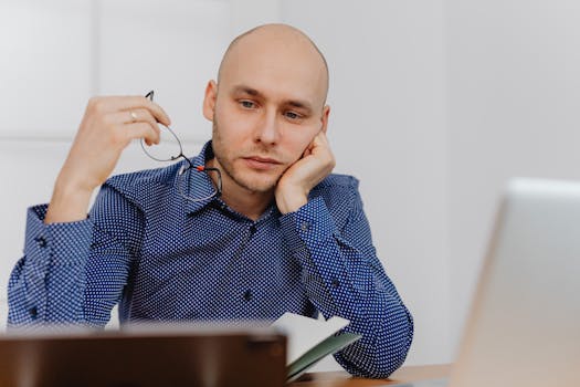 Bald businessman in blue shirt thinking at desk with glasses and laptop.