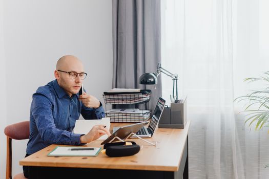 Bald man in glasses holding phone, sitting at a desk with laptop, in a home office.