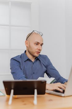 Bald man with eyeglasses working on laptop in a modern office setting.