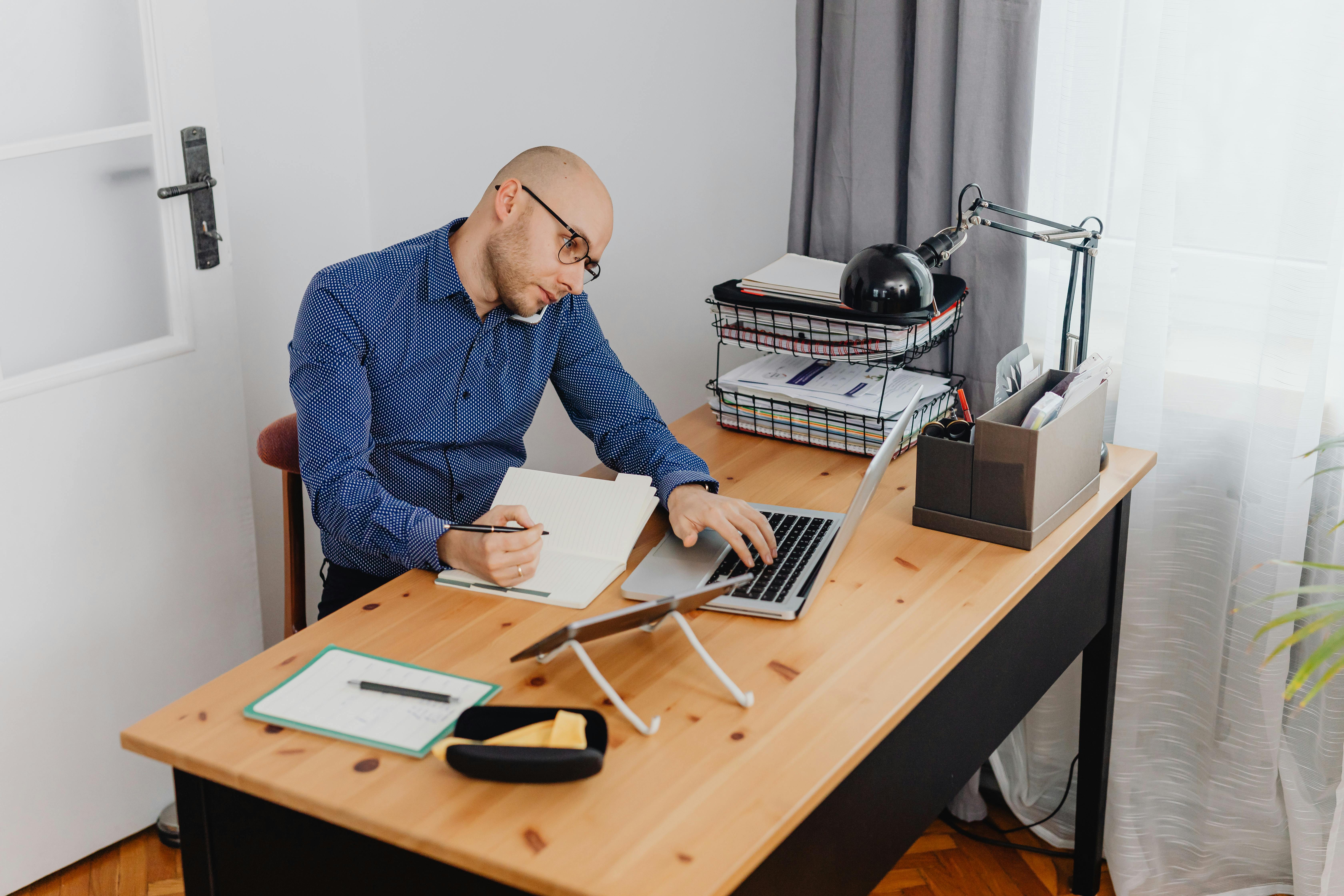 A man multitasks at his workspace, using a laptop and writing notes at a desk.