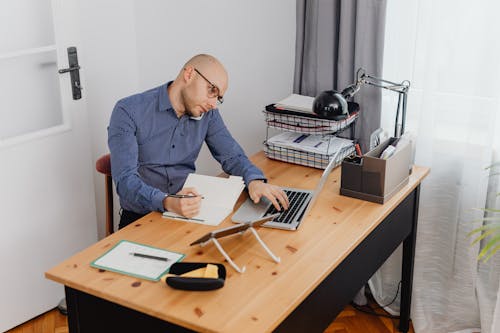 Free A Man on the Phone Holding a Pen Typing on a Laptop  Stock Photo