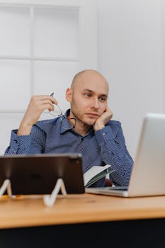 Thoughtful businessman holding glasses while working at his office desk using a laptop.
