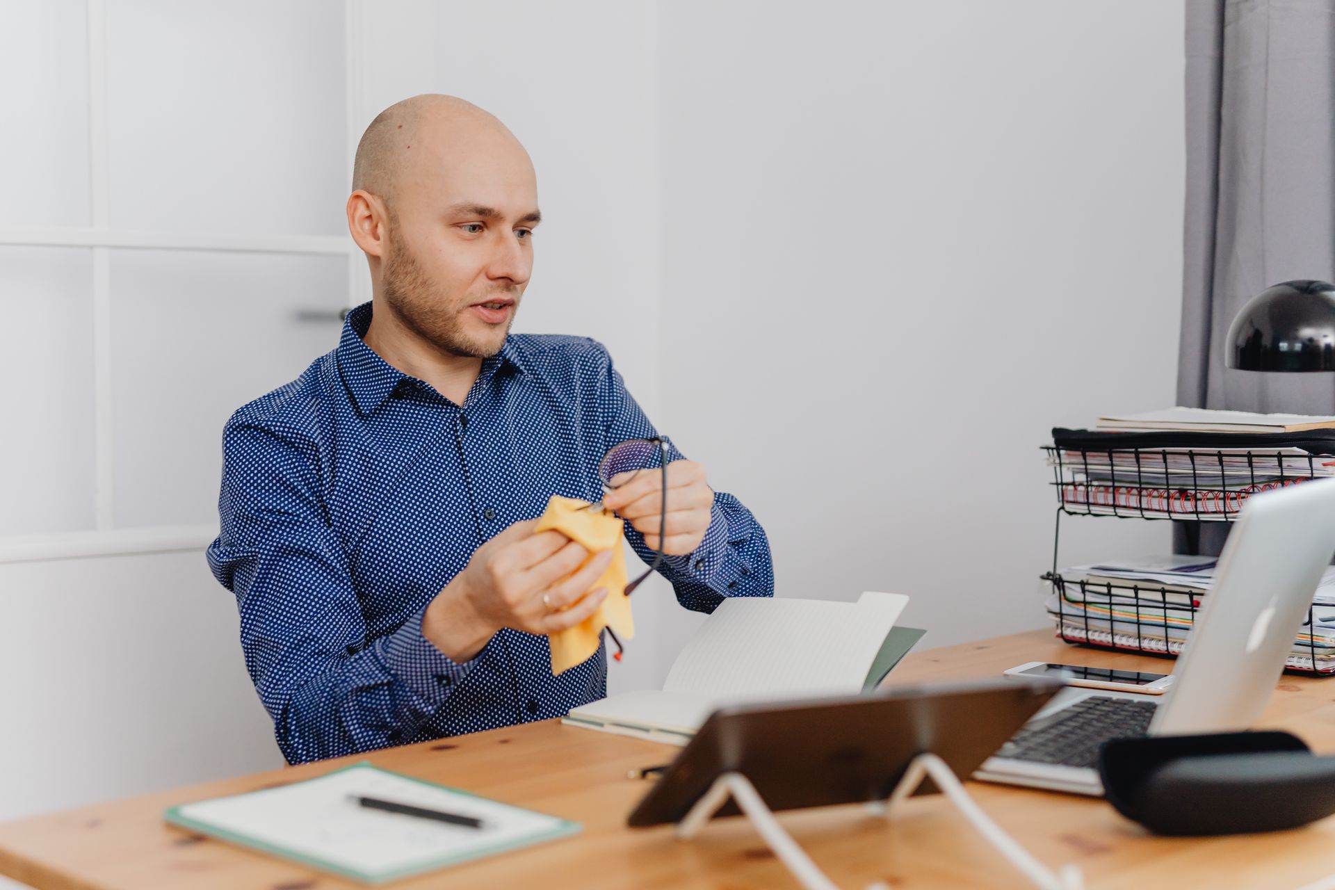 Bald man cleaning eyeglasses while working at an office desk.