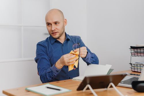 Free Bald man in a blue shirt cleaning eyeglasses at his office desk, emphasizing work setup and organization. Stock Photo