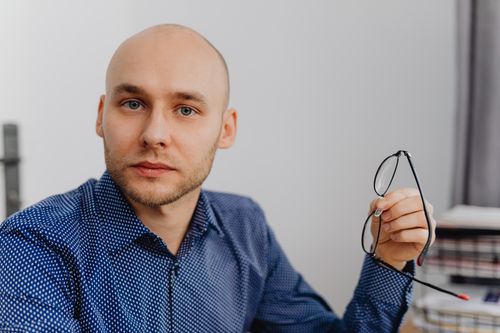 Free Bald man in a blue shirt holding eyeglasses indoors, with a thoughtful expression. Stock Photo