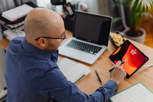 A man in an office setting using a laptop and tablet while taking notes.