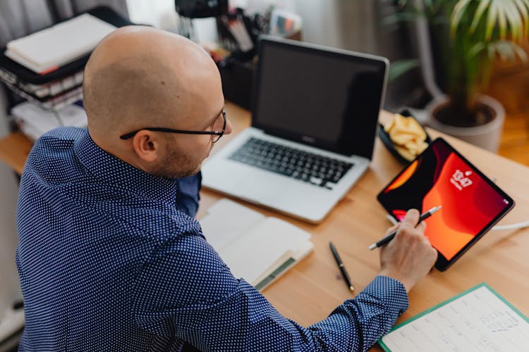 High Angle View Of A Balding Man In Glasses Working At Desk Using Devices